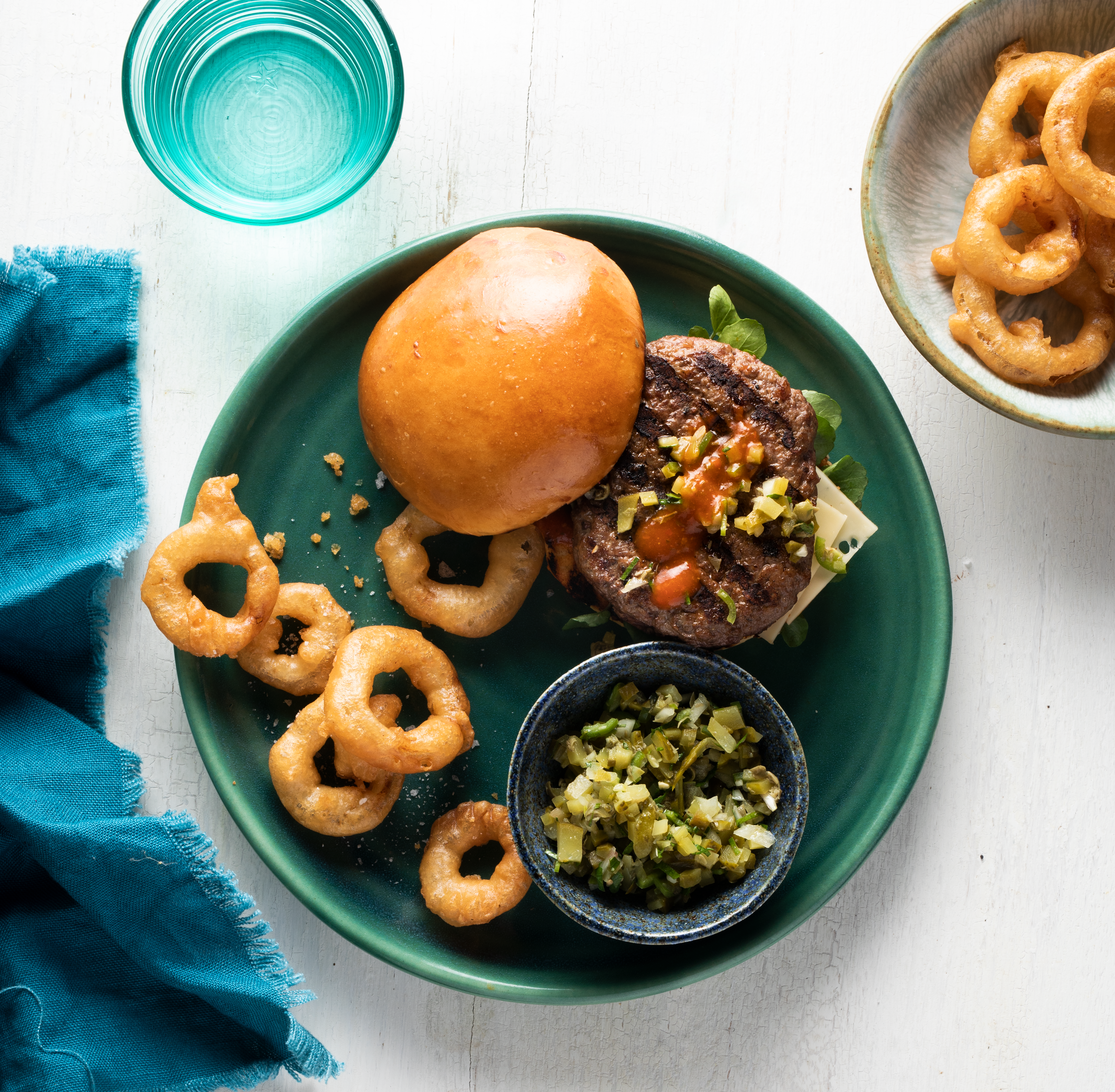 Beef and Lamb Burger with Gherkin Relish and Swiss Cheese on a green plate with fried onion rings and gherkin relish on the side
