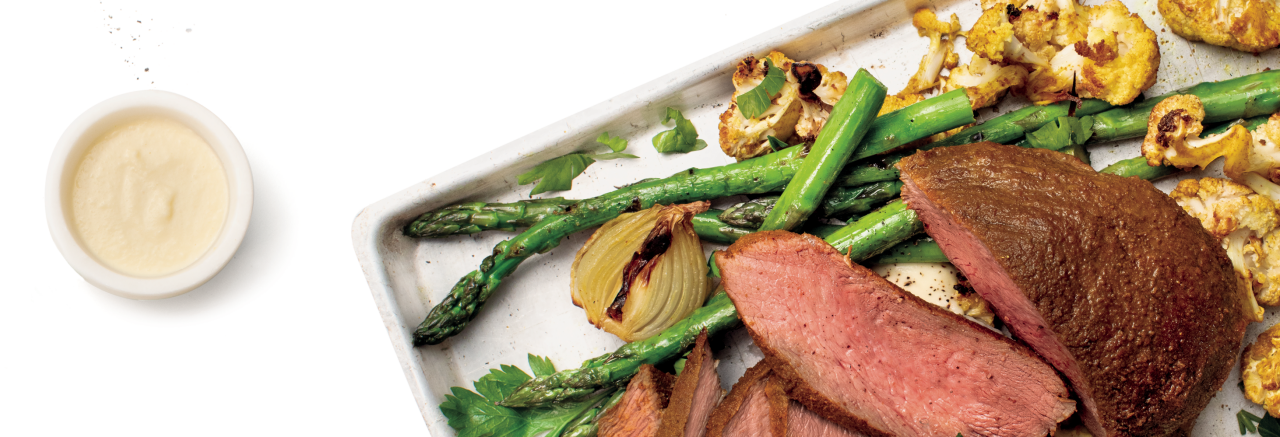 Venison Roast with Kale and Cauliflower and Turmeric Onions on an oven tray with a bowl of sauce