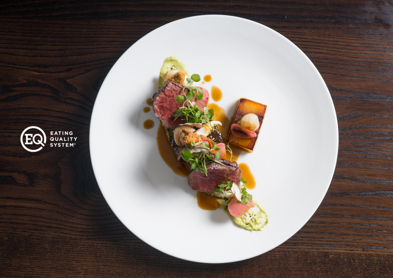 Beef entree with green toppings and sauce on a white plate, on top of a dark wooden table with Eating Quality System logo.