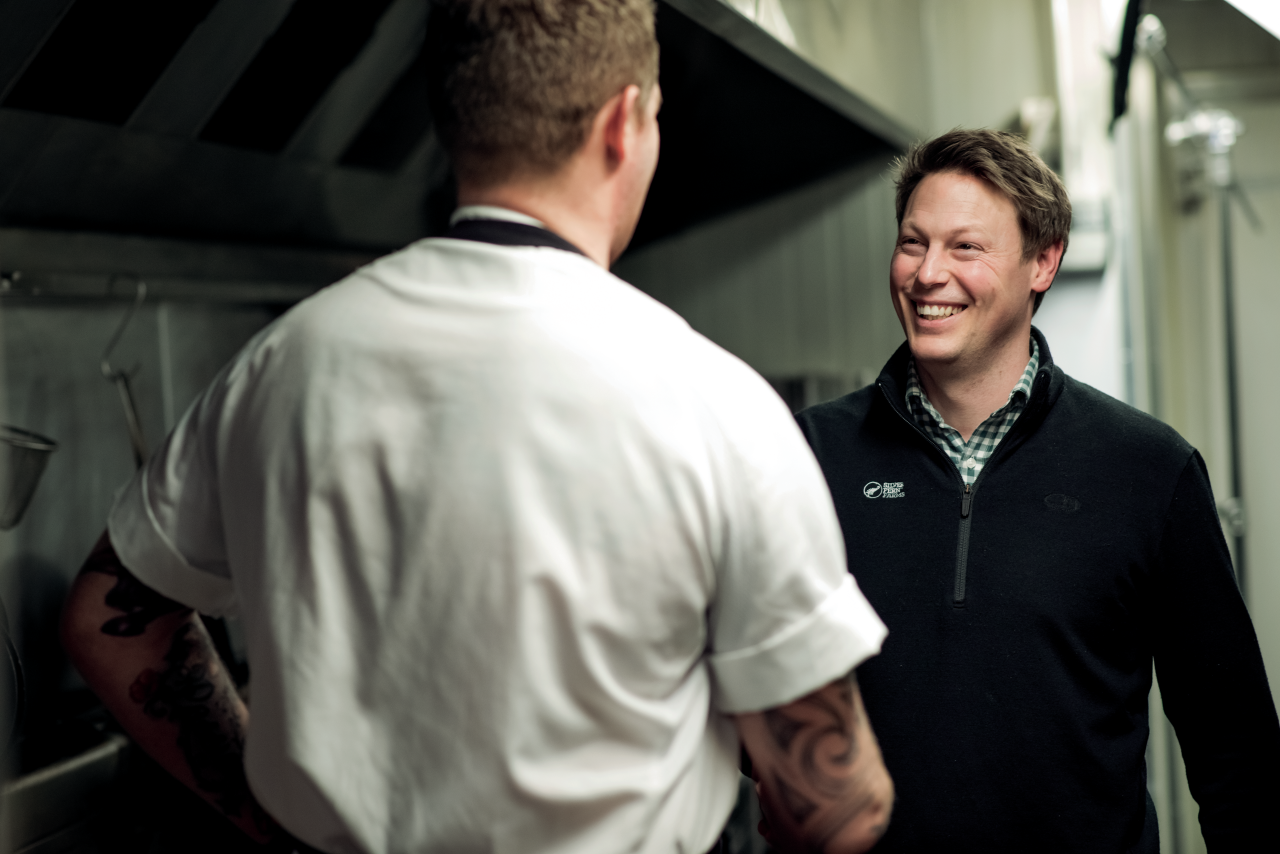 Silver Fern Farms staff member smiling while speaking to a person in chefs uniform, in the kitchen of a restaurant.