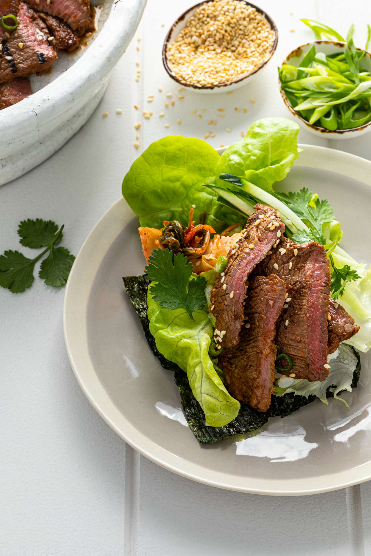 Korean Inspired Venison Minute Steaks Lettuce Cups with Kimchi, Rice and Cucumber on a cream plate with a bowl of sesame seeds and spring onion for garnish on the side