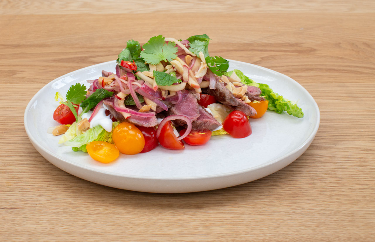 Venison steak, Thai style apple salad with yoghurt and peanuts on a white plate on a light wood bench