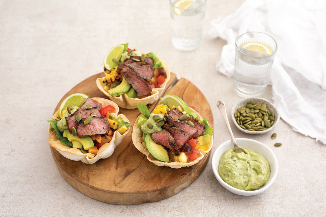 Venison Medallions Taco Salad Bowls with Avocado and Corn on a wooden chopping board with guacamole and pumpkin seeds as side dishes