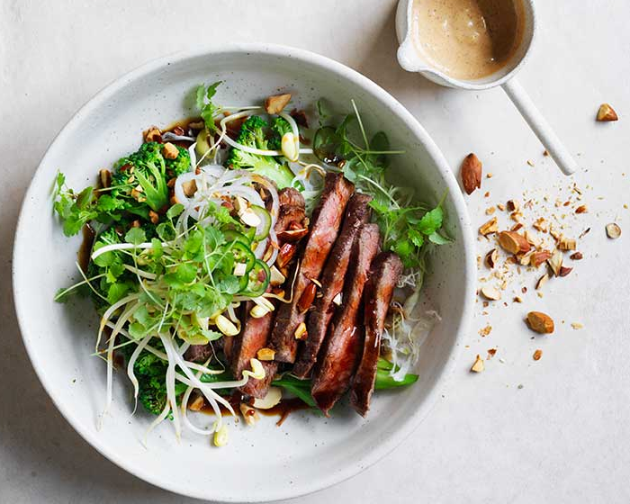 Warm Broccolini and Steak Salad with Almond Satay Sauce in a white bowl next to crushed almonds